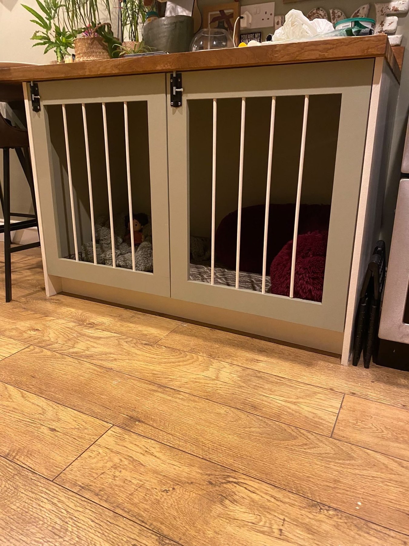 Two dog crates built into the end of a kitchen island, with sage green shaker doors, white spindles and a solid oak worktop over