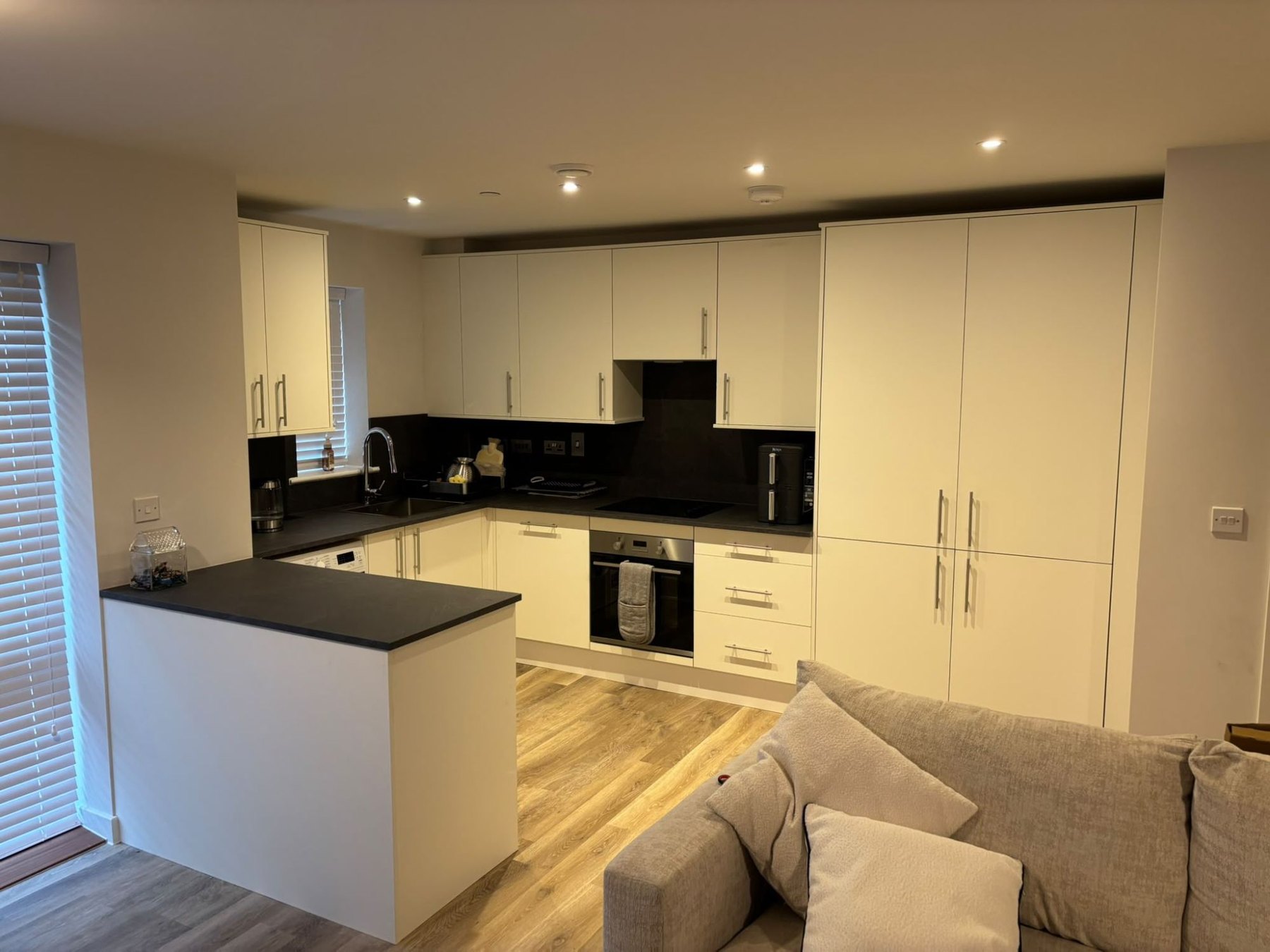 Cream fitted kitchen with a peninsula worktop dividing it from the living room, dark worktops and full-height cupboards