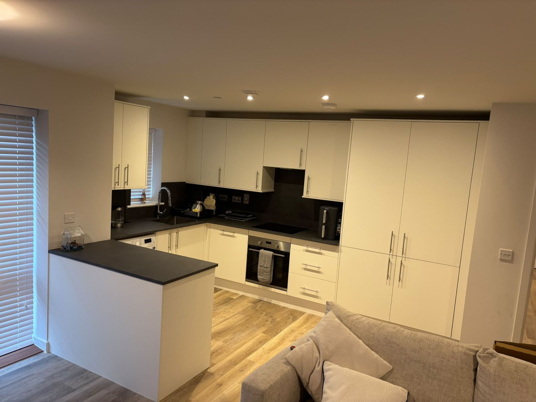 Cream fitted kitchen with flat slab doors fitted around the corner of a room, with dark worktops, a run of tall units and an integrated oven