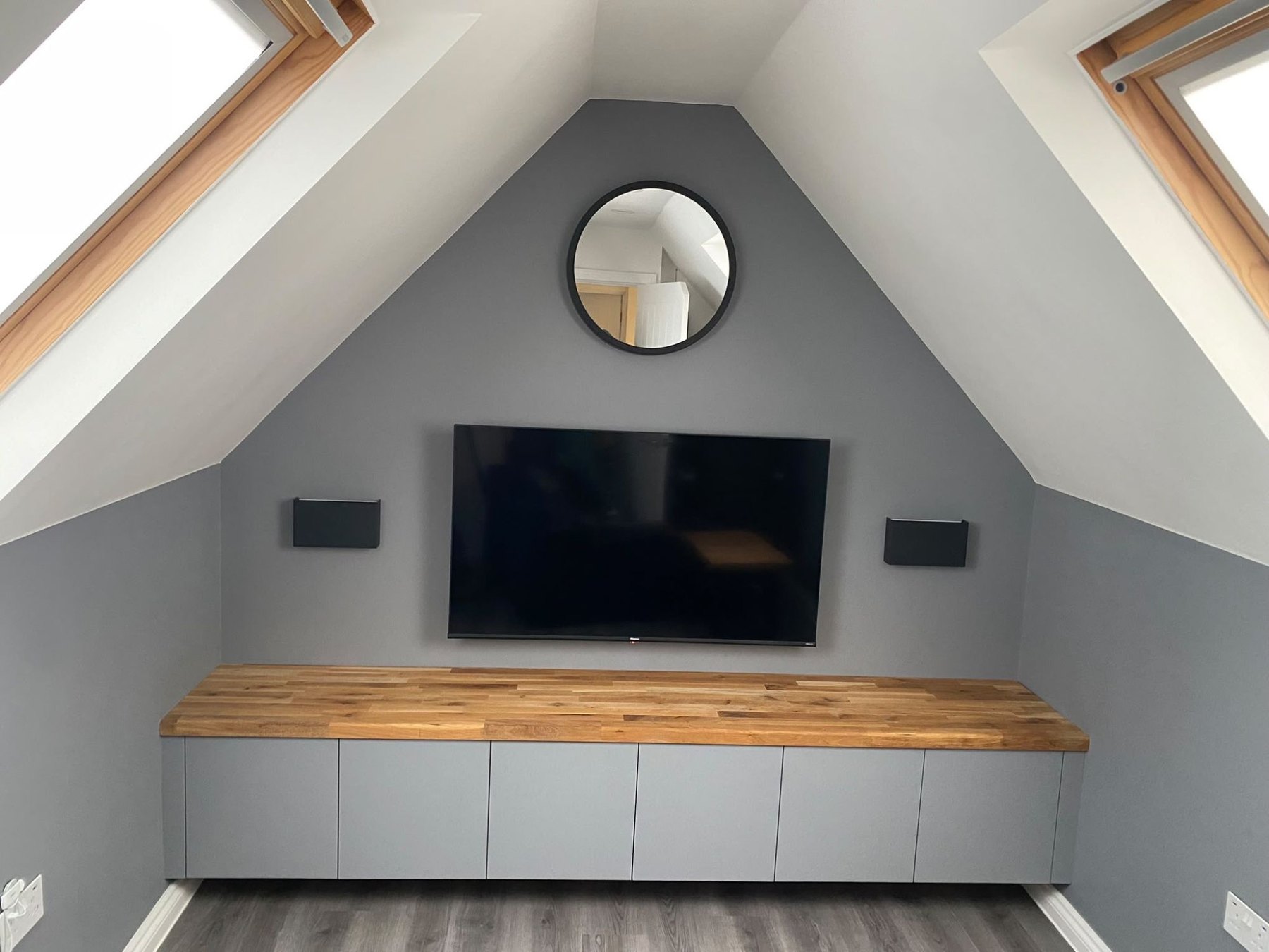 Low fitted unit running the width of a loft room under the sloping ceiling, with a solid oak worktop and a wall-mounted television above