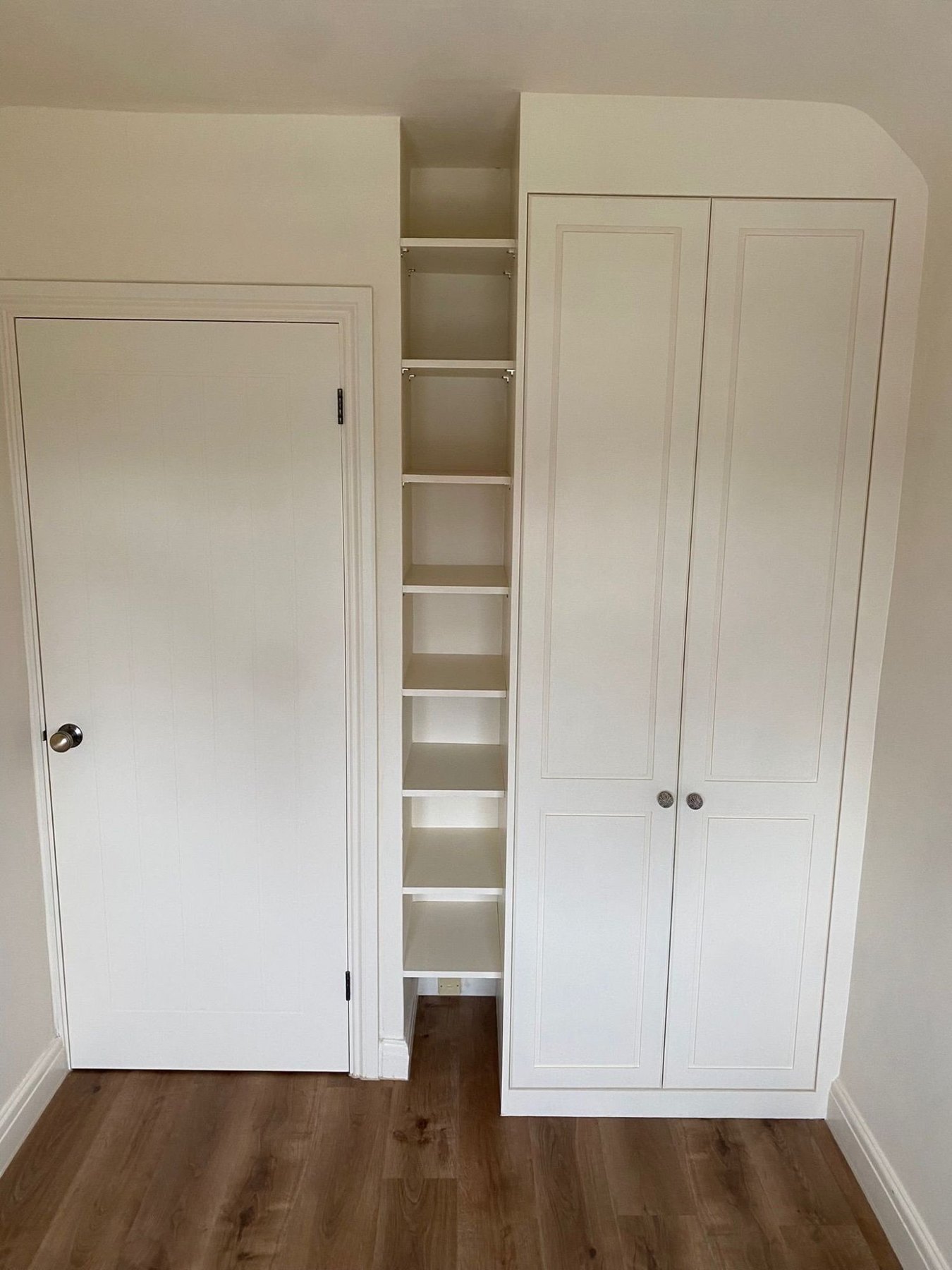 Painted wardrobe with panelled doors and a rounded top corner, beside a column of open shelving fitted into the recess